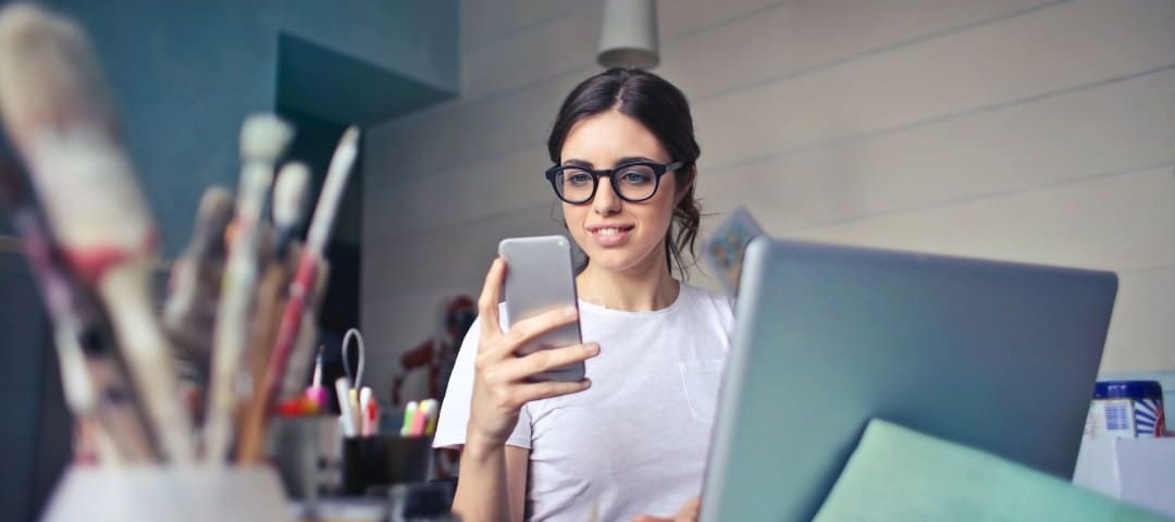 Photo of a woman in front of a computer looking at her phone