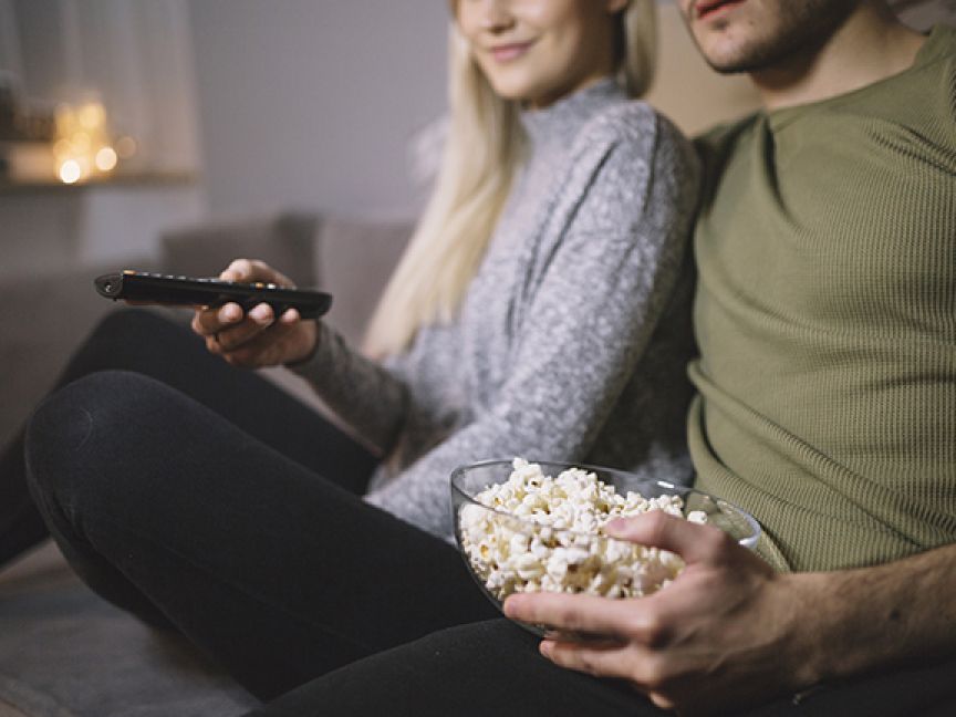 Two people sit on a couch with popcorn and a remote control
